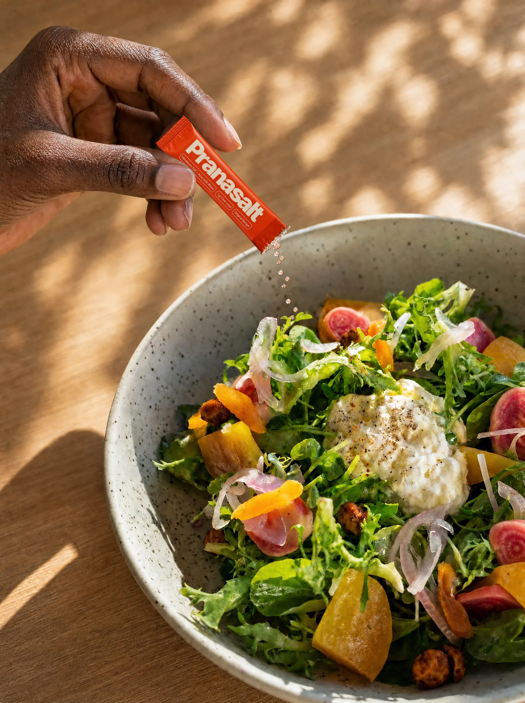 A hand sprinkling a Pranasalt sachet over a finished salad