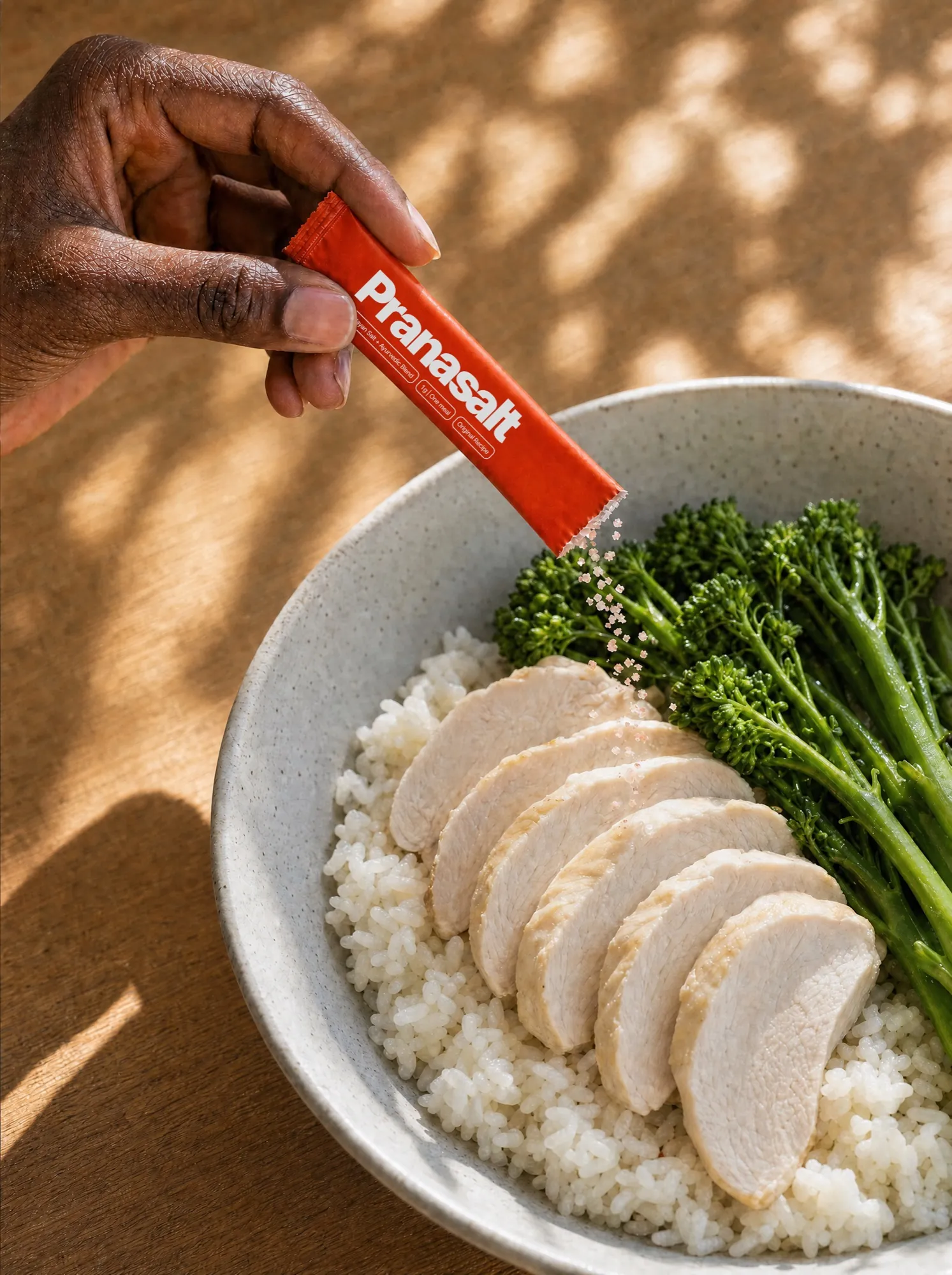 A hand sprinkling a Pranasalt sachet over a bowl of chicken, rice and broccolini