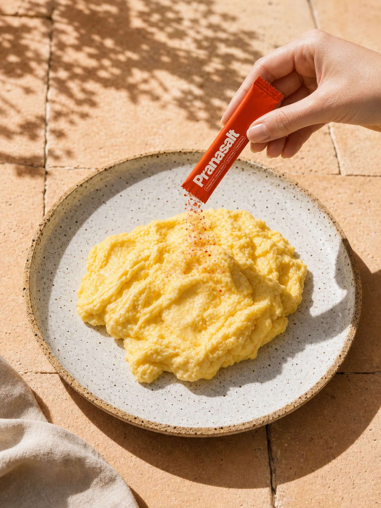 A hand sprinkling a Pranasalt sachet over a plate of scrambled eggs in warm sun