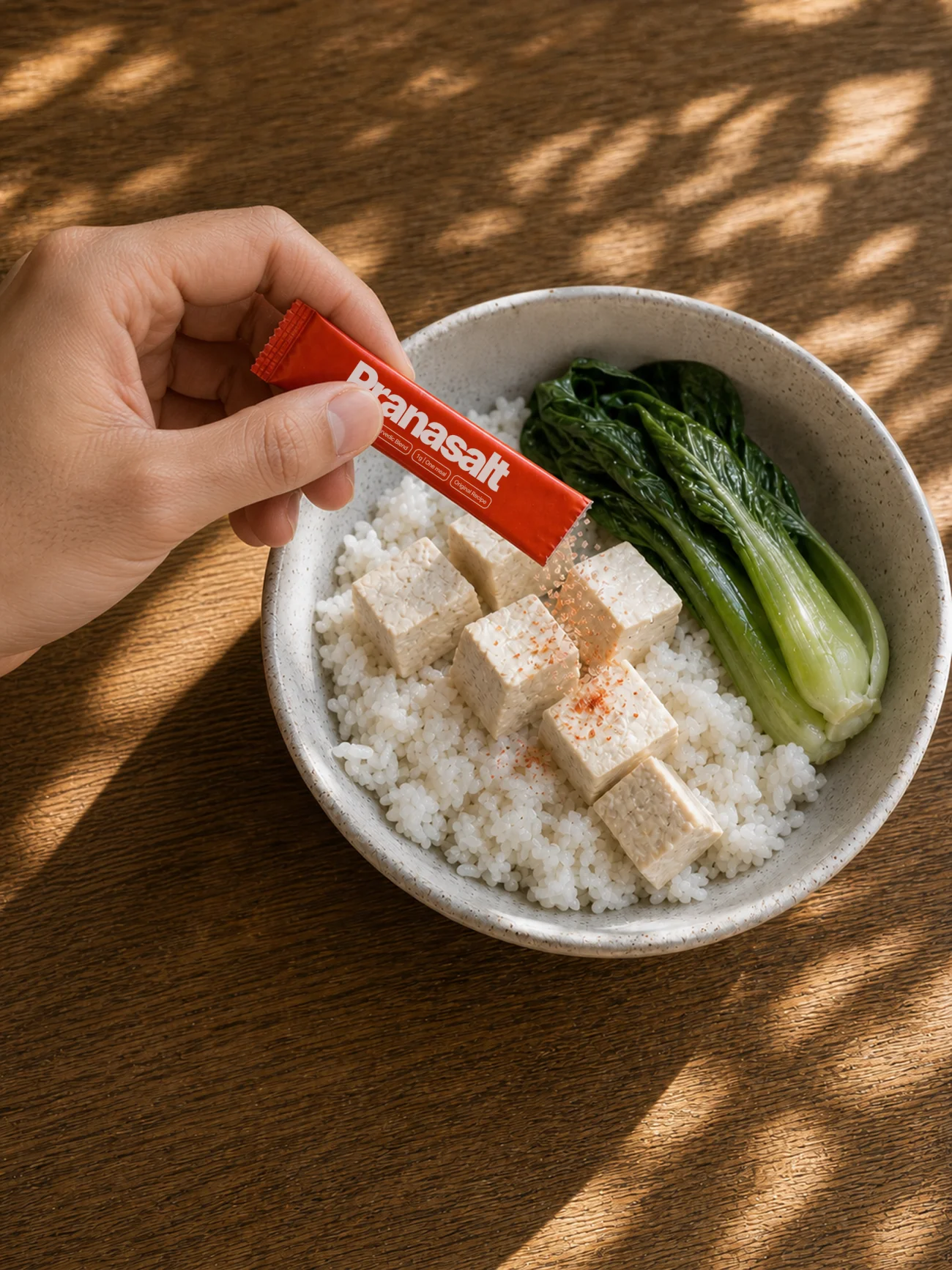 A hand sprinkling a Pranasalt sachet over a bowl of rice, tofu and bok choy